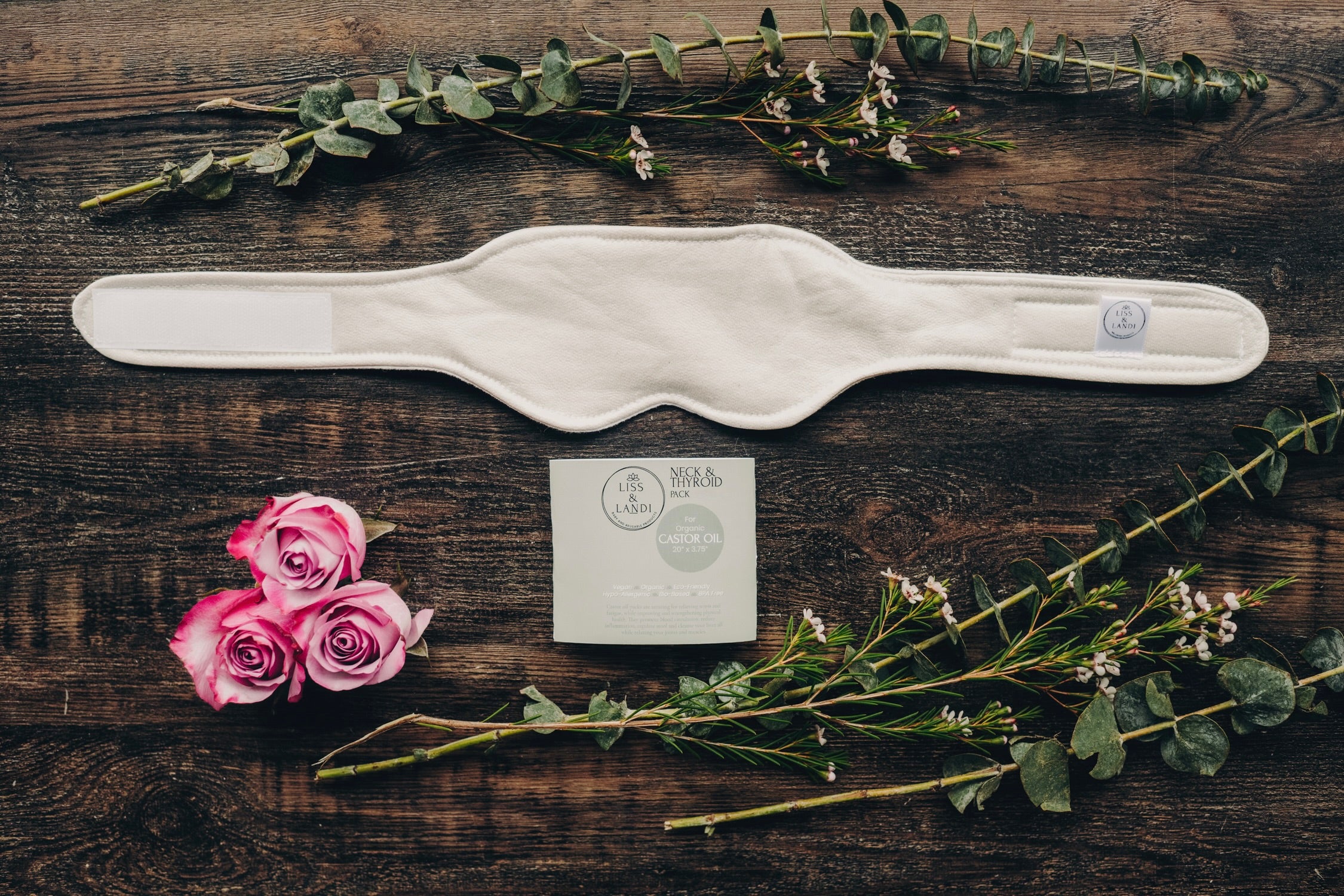 A beige neck and thyroid organic castor oil pack laid out flat with a piece of flannel, surrounded by decorative flowers and greenery.