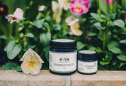Two jars of comfrey salve on a wooden surface with flowers in the background