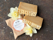 A bar of artisan soap with a honeycomb pattern on top, wrapped in brown paper with a label, accompanied by yellow flowers.