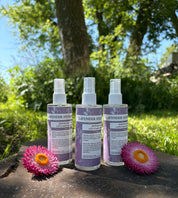 Three bottles of Lavender Hydrosol products on a wooden surface with flowers and greenery in the background.